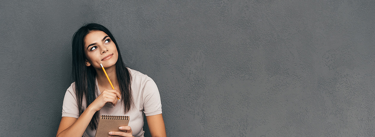 Attractive young woman in casual wear holding notebook and pen while sitting barefoot and against grey background